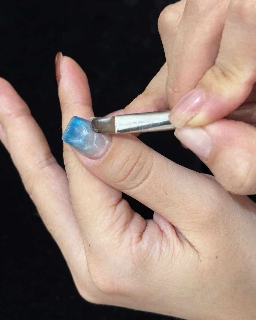 A brush dabbing blue glass gel on the tip of a manicured nail to create an ombre gradient.