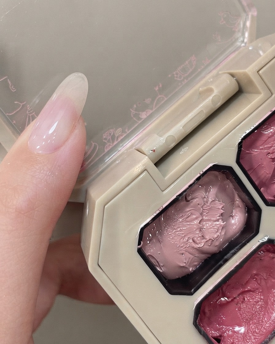 A sponge applicator hovering over a pink pigment palette next to a hand with a natural nail.