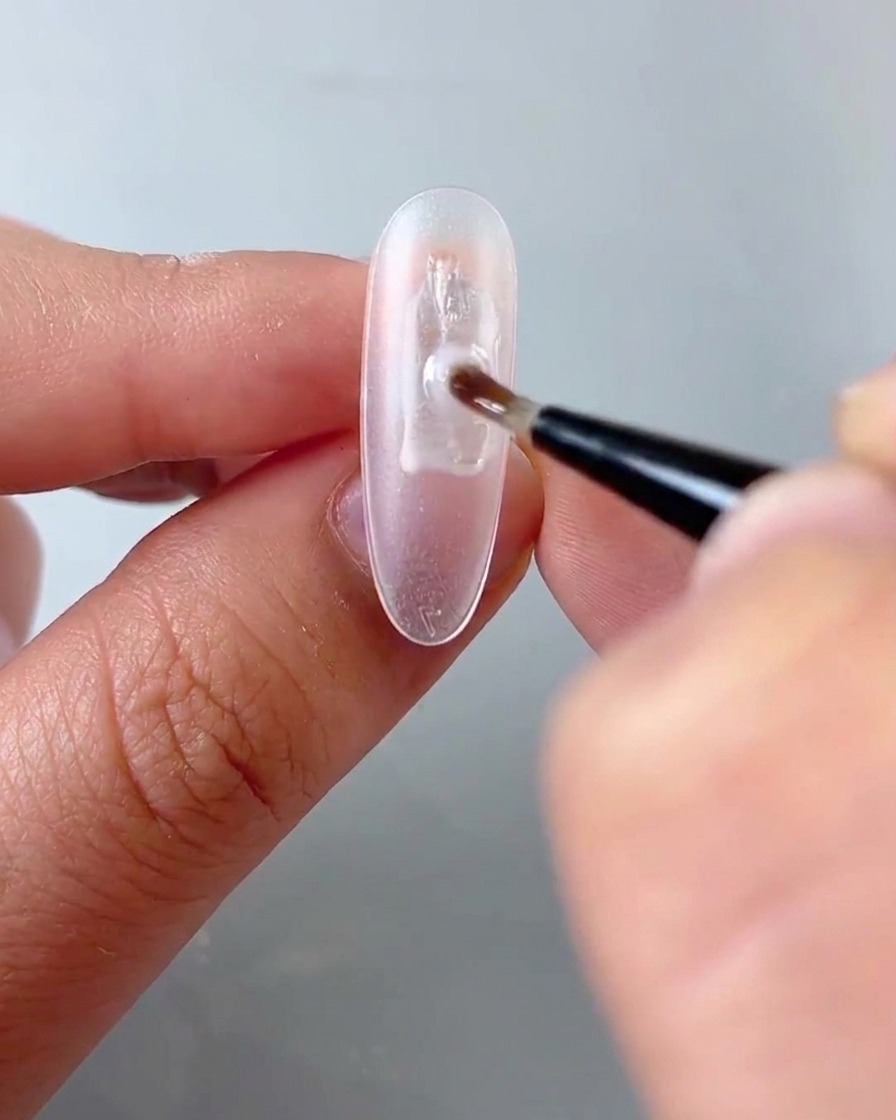 A close-up view of a fine brush applying clear gel onto an oval nail plate.