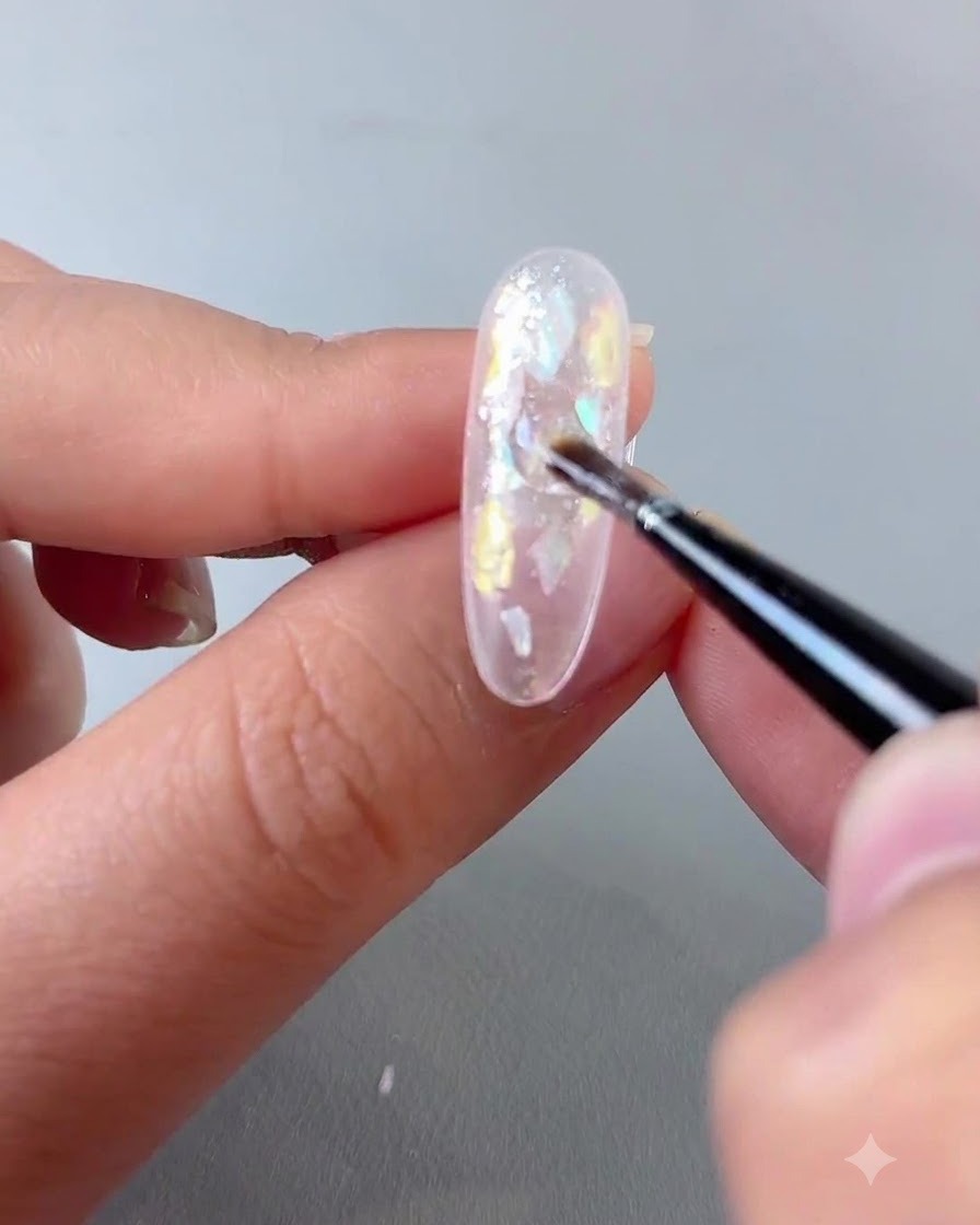A brush applying a sparkly gel mixture evenly over the decorated surface of a nail.