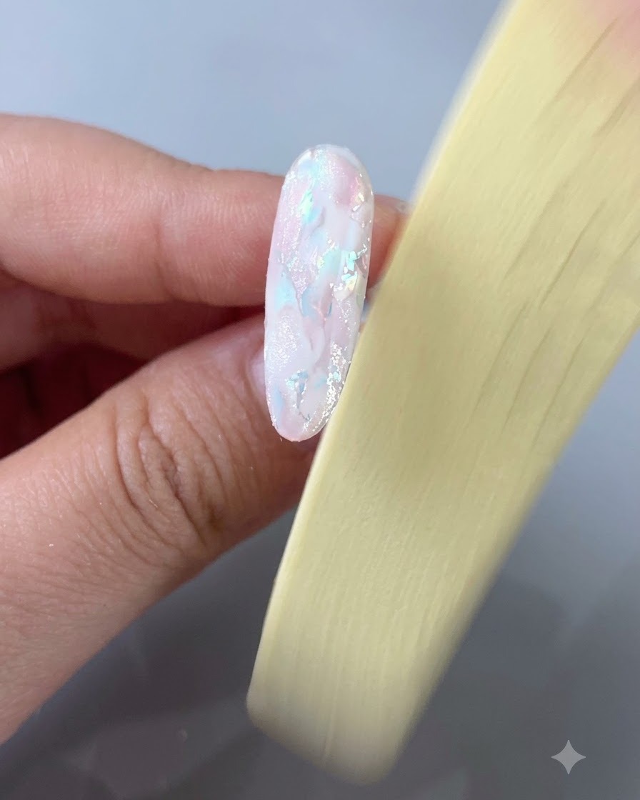 A close-up view of a hand using a yellow nail file to buff the surface of an almond-shaped nail featuring a milky white base and iridescent shell pieces.