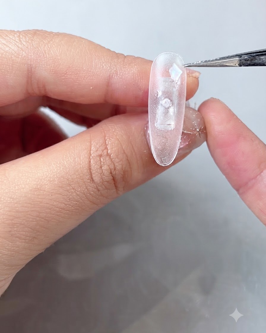 A close-up of tweezers placing iridescent blue shell pieces onto a clear, frosted nail tip coated with wet fixing gel.