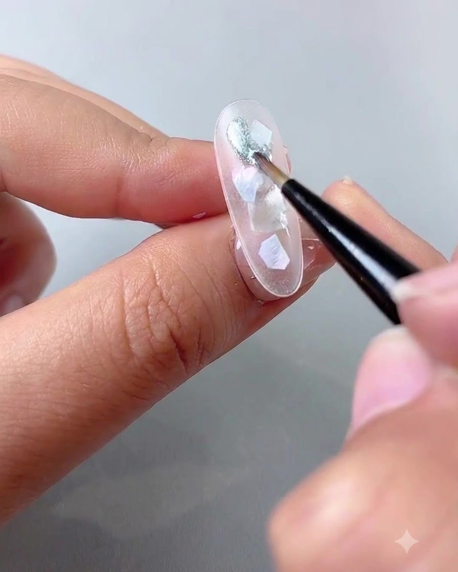 A thin nail art brush applying a layer of sparkly, grayish-blue cat eye gel over iridescent shell pieces on a clear nail.