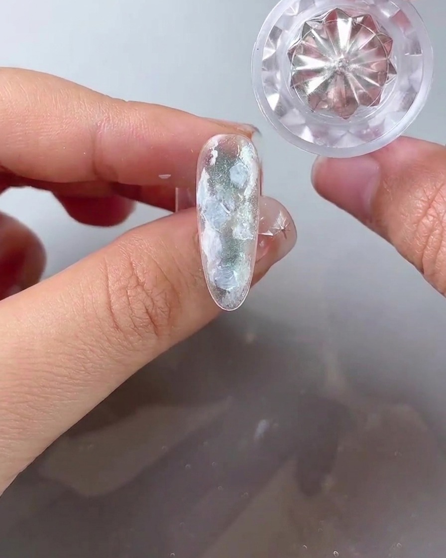 A fine liner brush dropping milky white gel into the gaps of a shell and pearl nail design.