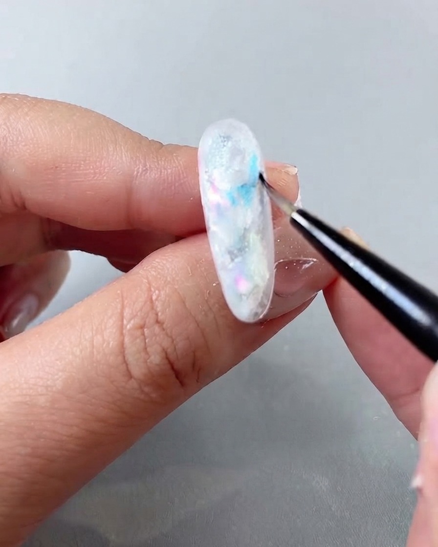 A fine nail art brush applying translucent blue glass gel into the gaps of a marble nail design.