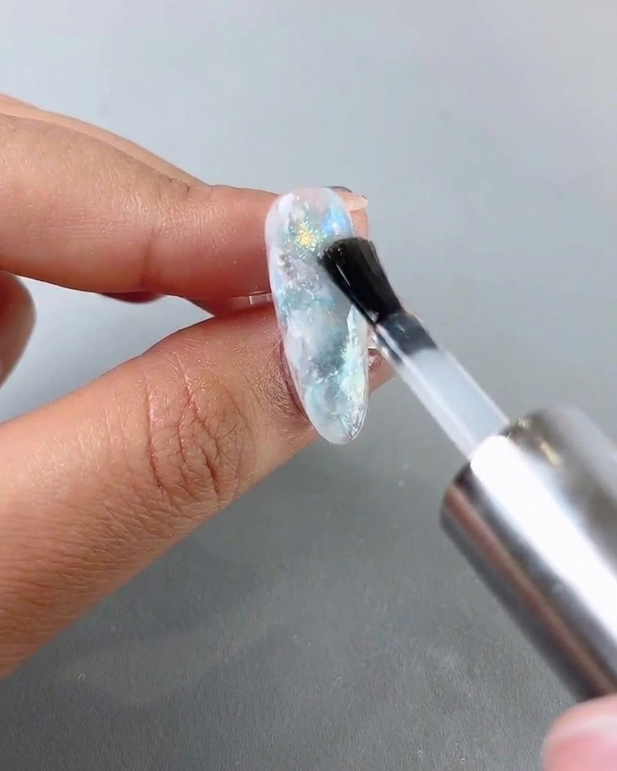 A brush applying a glossy top coat layer over a completed blue and white marble nail.