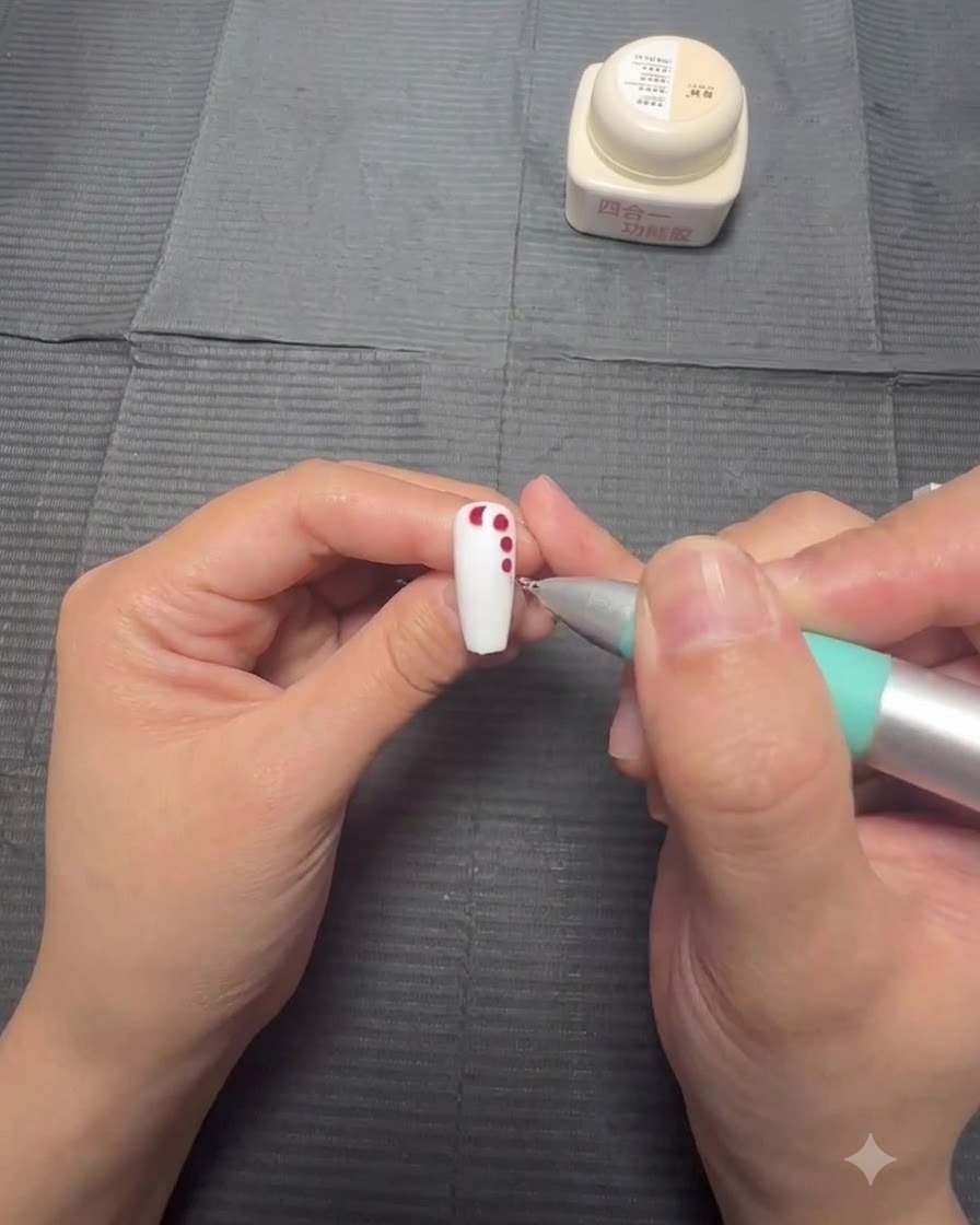A hand holding a nail while a dotting pen applies progressively smaller red gel dots in a downward line over a white base.