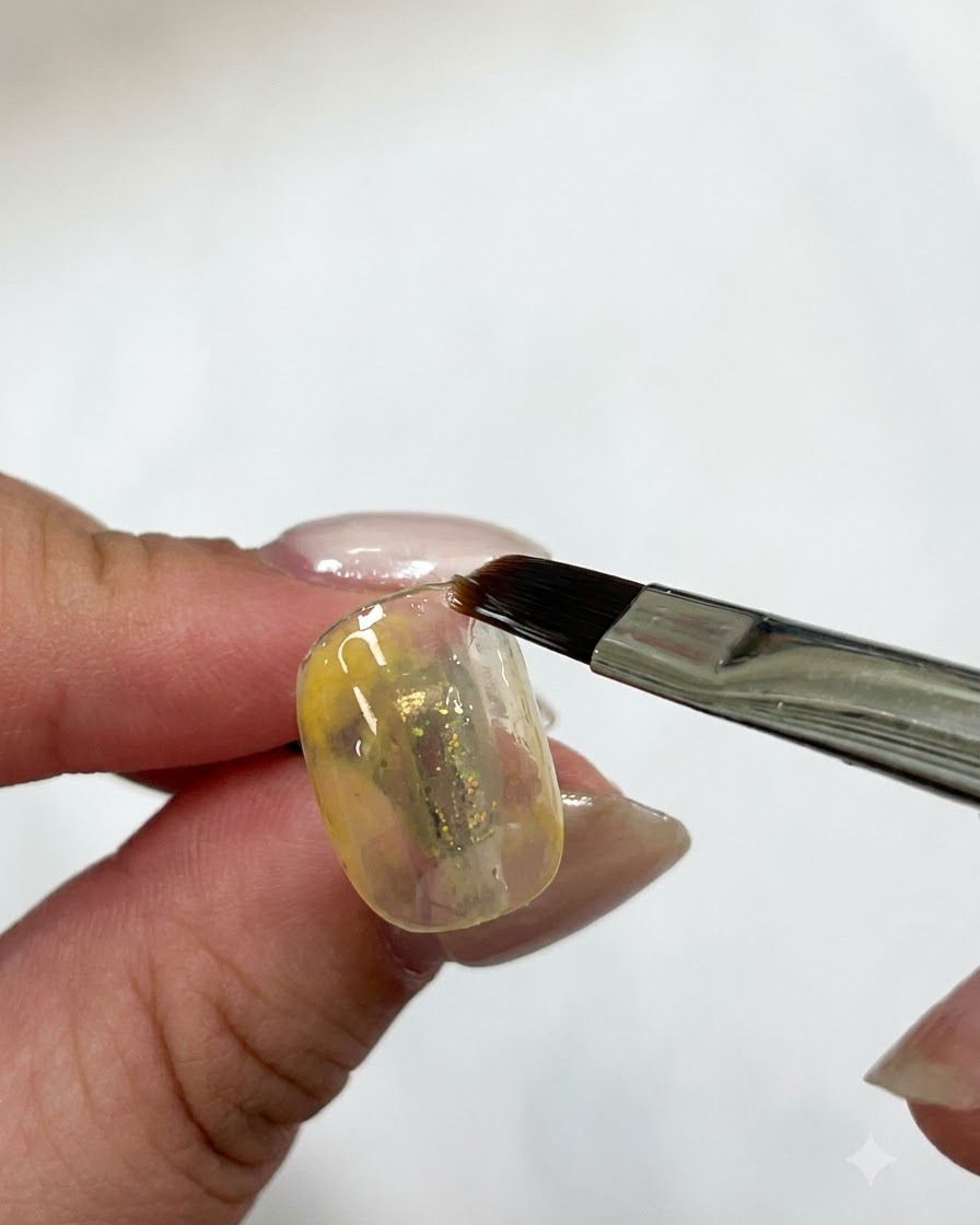 A flat gel brush smoothing the yellow gel layer on a nail under lighting.