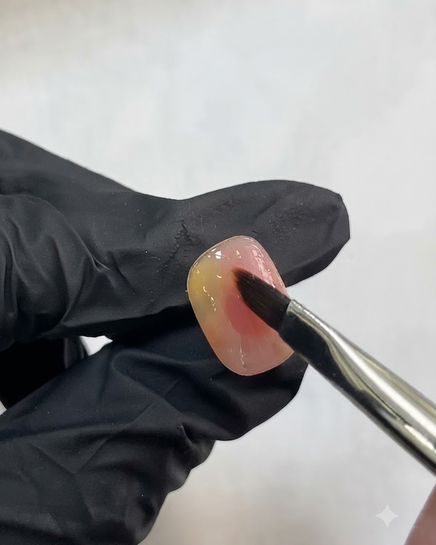 A nail brush delicately applying a small stroke of translucent white gel onto a pink watercolor nail.