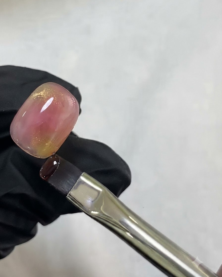 A fine liner brush holding a drop of gold cat eye gel near the top of a pink watercolor nail.