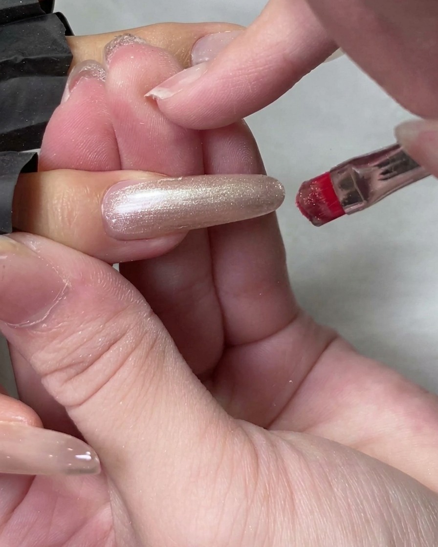 A bottle brush applying translucent cat eye gel to a long natural fingernail.
