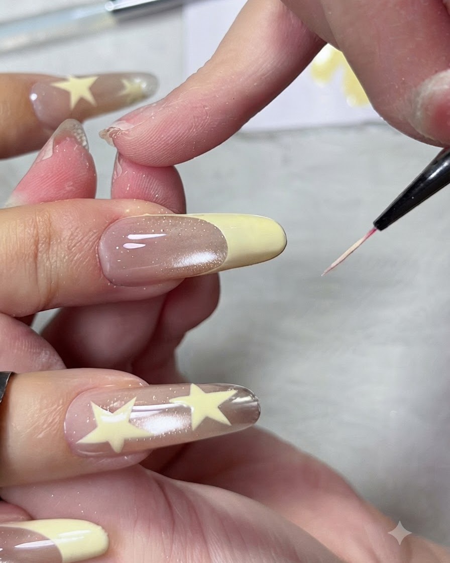 A close-up view of a fine liner brush shaping and sharpening the smile line of a pastel yellow French manicure tip on a long almond nail.