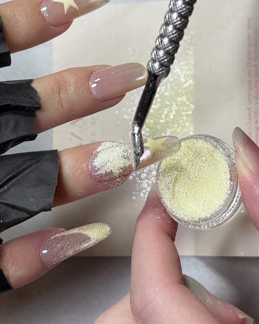 A metal cuticle pusher scooping and showering sparkly creamy yellow crushed diamond powder over wet painted star designs on a fingernail.