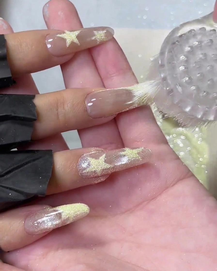 A round plastic dusting brush sweeping loose yellow powder off long nude acrylic nails with textured star details.