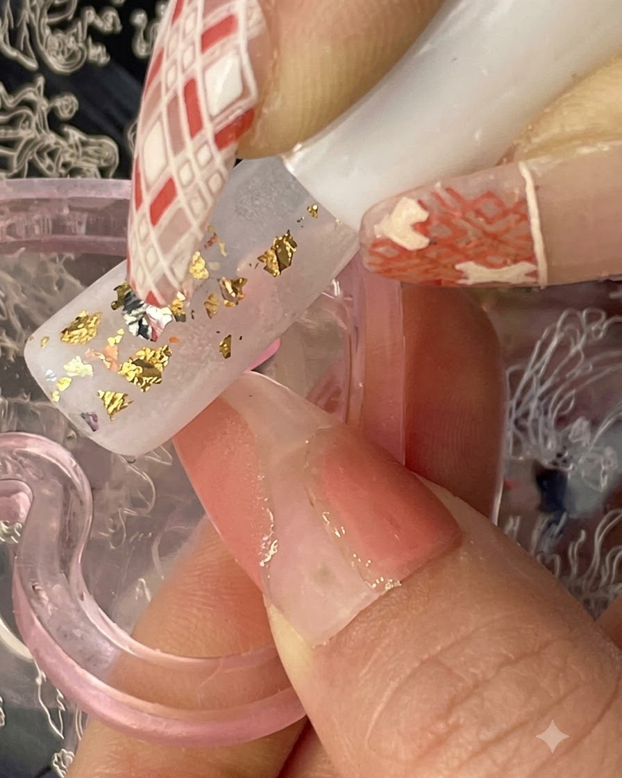 A nail brush applying translucent brownish-orange iced tea gel in irregular patches over a base with gold flakes.