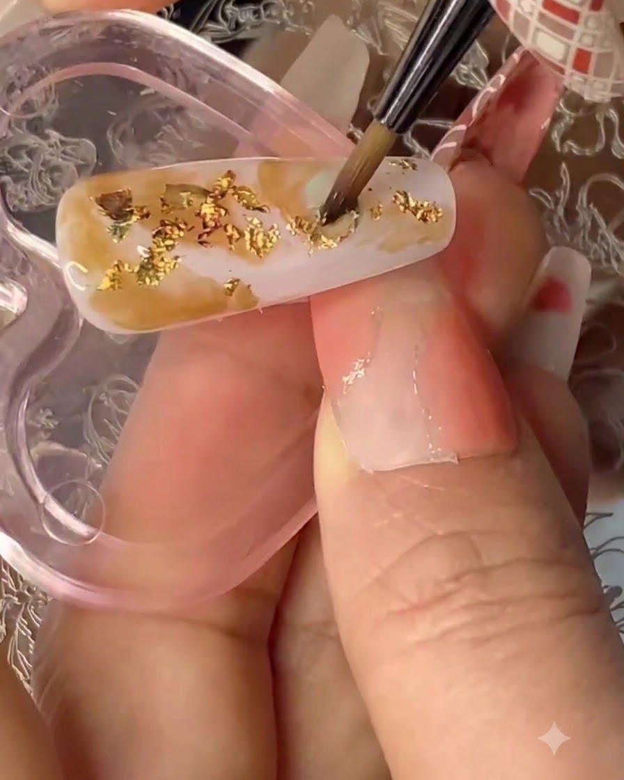 A fine detail brush applying clear solid gel over a textured nail with gold flakes and smudged colors.