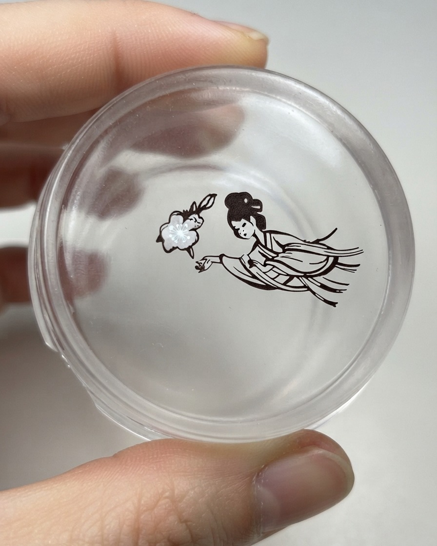 A clear silicone stamper head holding a transferred design of a flower next to a detailed traditional female figure.