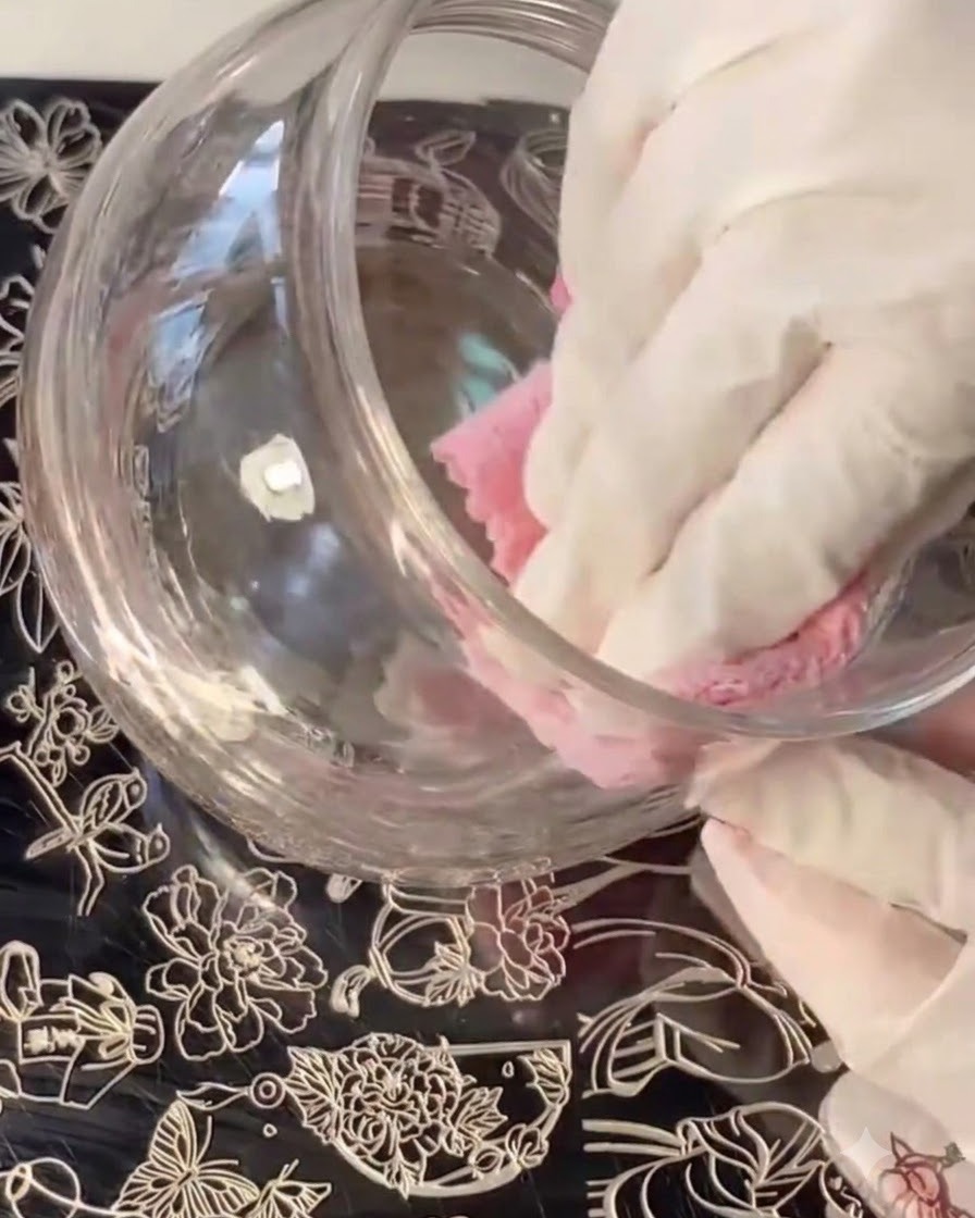 A gloved hand cleaning or wiping a glass container while stamping plates with floral designs lie underneath.