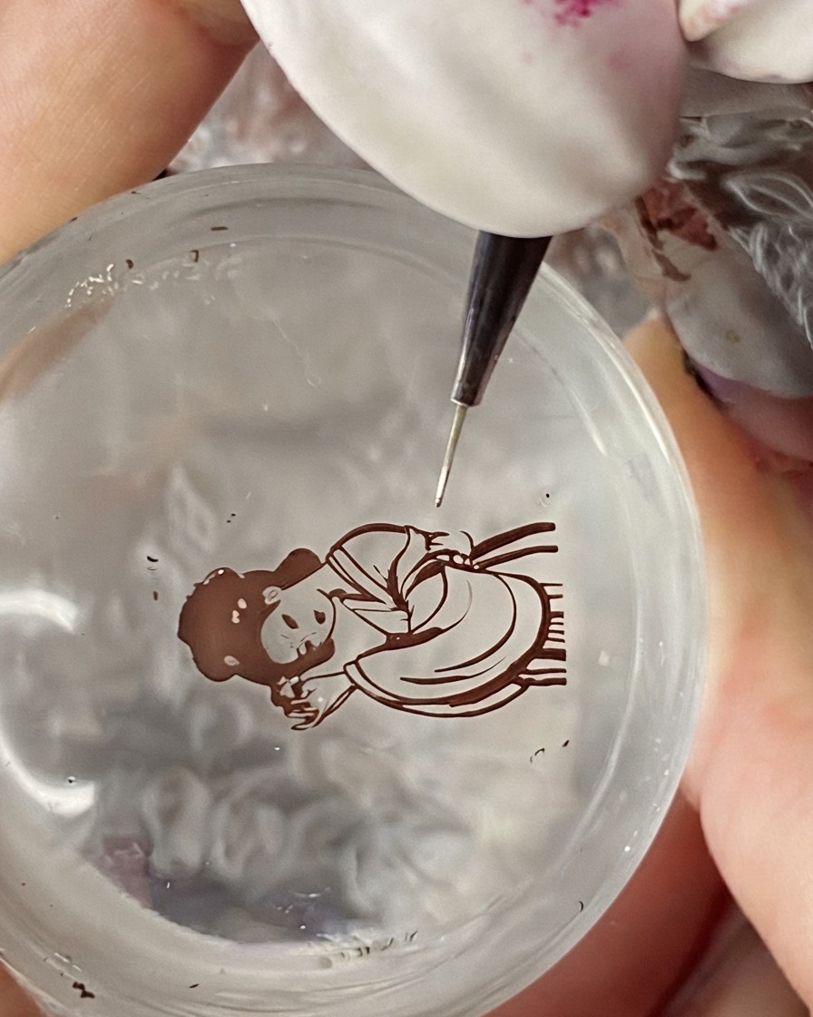 A fine detail brush applying color to the clothing ribbon of a stamped traditional Chinese character design.