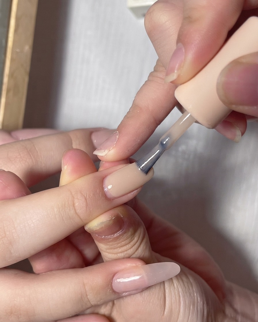 A polish brush applying a milky lighter gel color onto the free edge of a nude pink painted nail.
