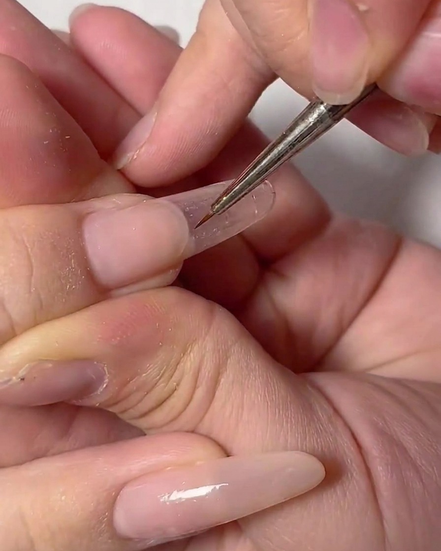 A close-up of a hand using a fine nail art brush to draw a delicate white outline on a clear nail.