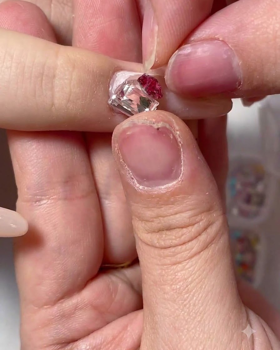 A pair of tweezers placing a large square crystal and a red floral rhinestone cluster onto a decorated nail.