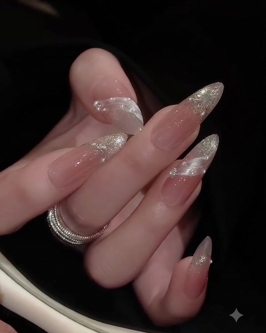 A close-up of a hand with almond-shaped nails, displaying clear glossy top coat and embedded rhinestones under soft lighting.